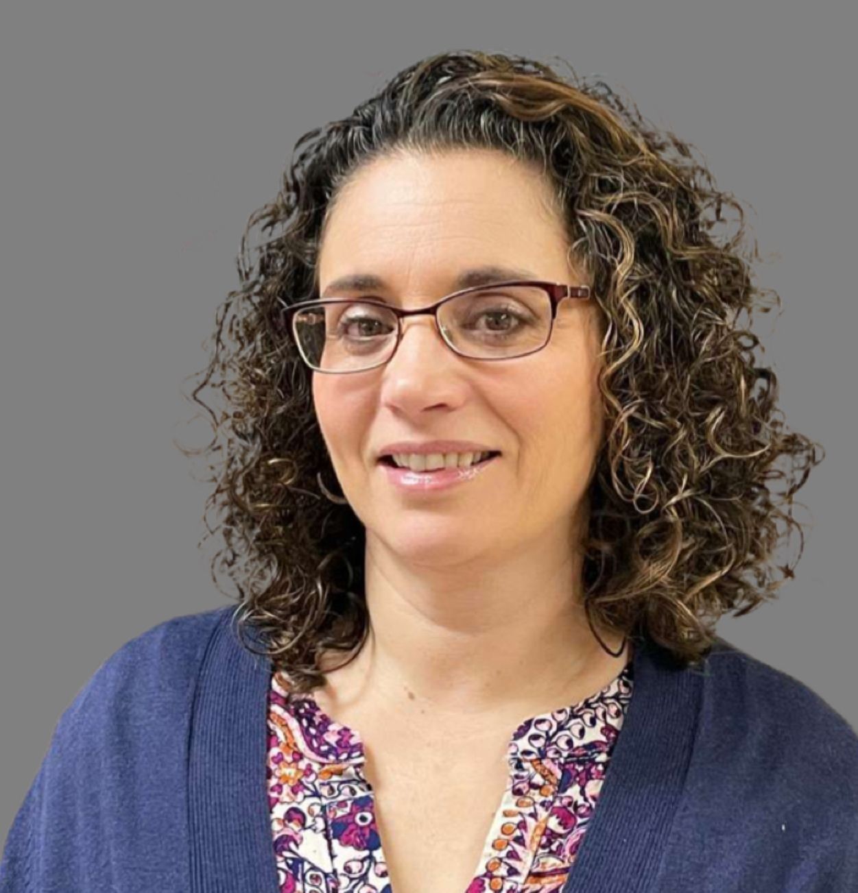 Woman with curly hair and glasses wearing a colorful blouse and blue cardigan against a gray background.