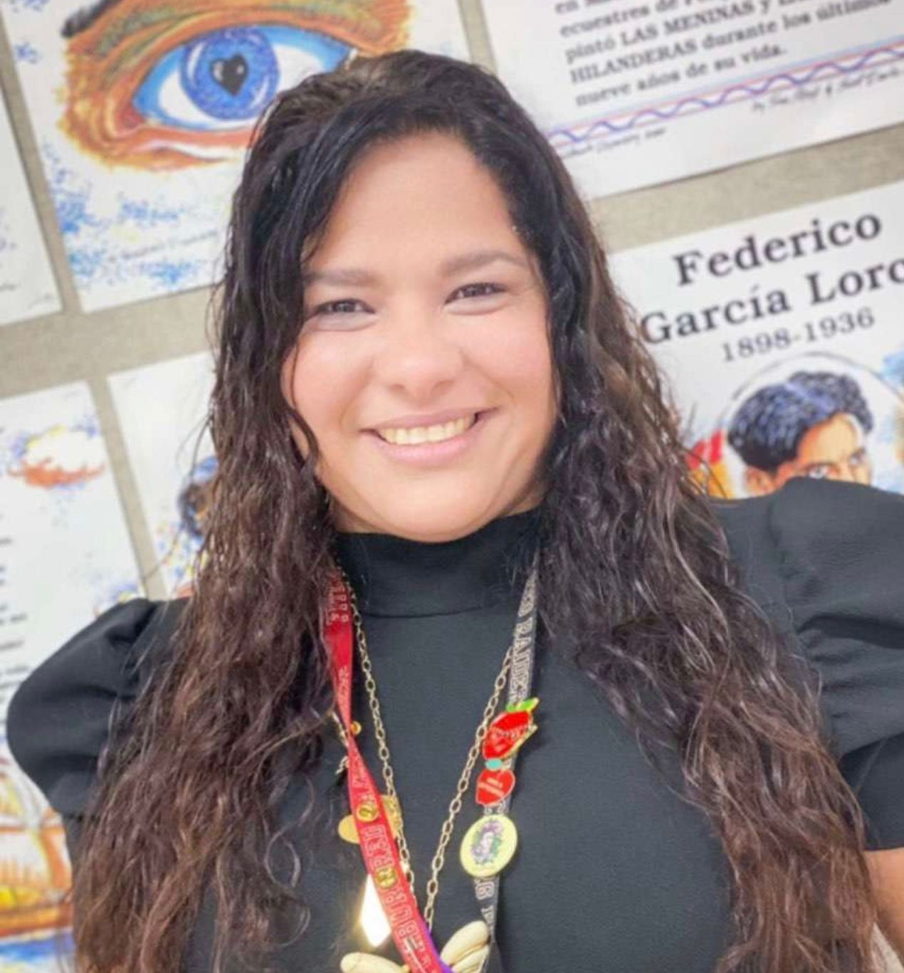 Smiling woman in black top with lanyards, standing in front of colorful posters.