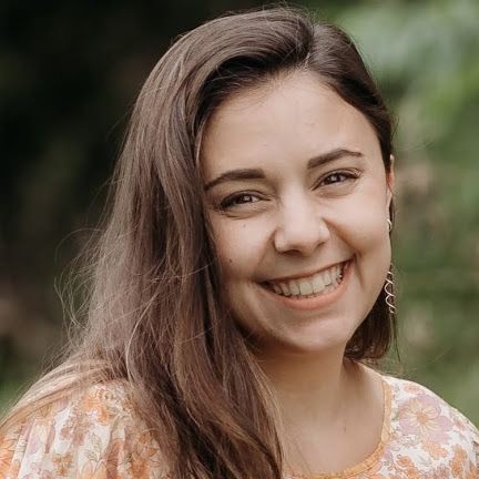 Person with long brown hair wearing a patterned blouse, standing outdoors with greenery in the background.