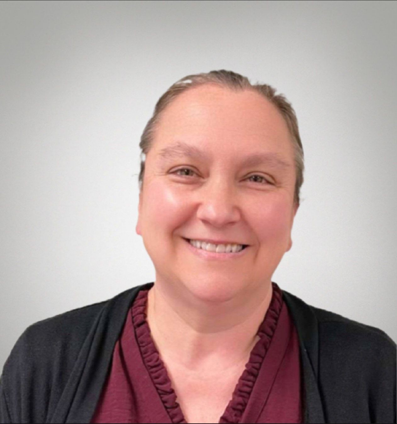 Smiling person with short hair wearing a maroon top and black cardigan against a gray background.