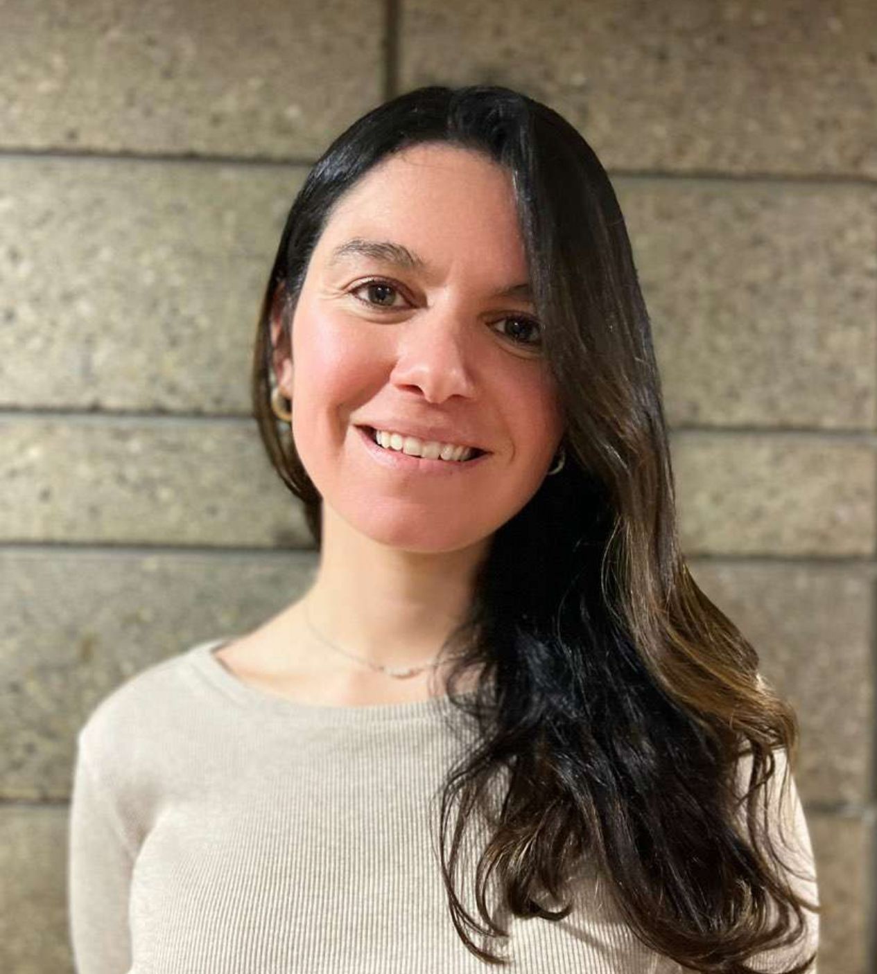 Person with long dark hair wearing a beige ribbed sweater standing against a textured wall.